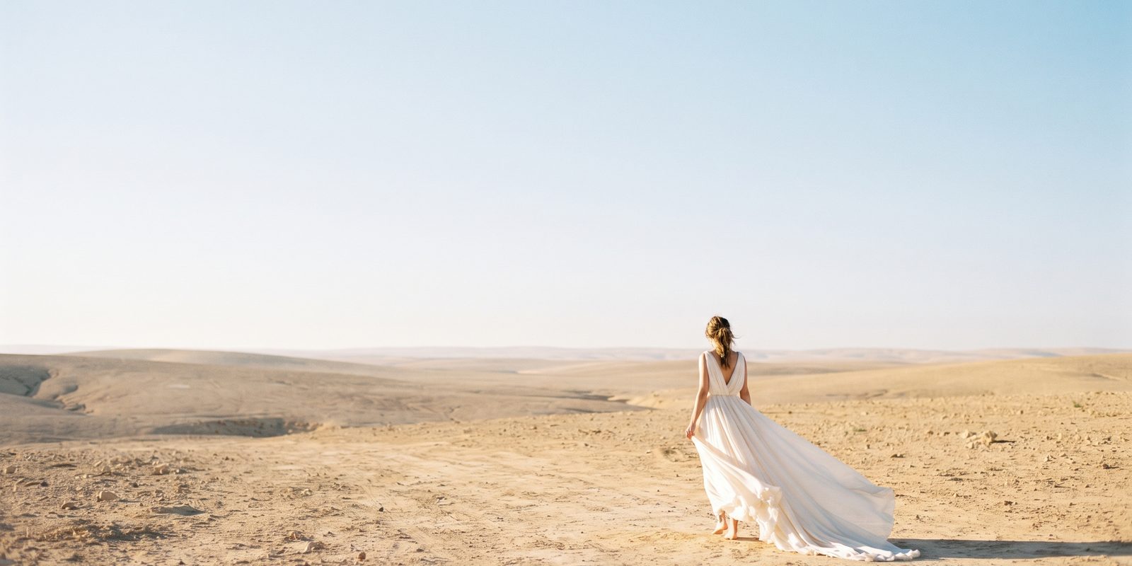 Frau in einem hellen Kleid mit langer Schleppe in einer weiten, leeren Landschaft